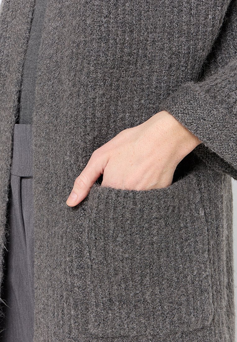 Grauer Strickcardigan mit strukturiertem Stoff, ausgestattet mit einer Fronttasche. Das Design umfasst einen lässigen Schnitt und weite Ärmel.