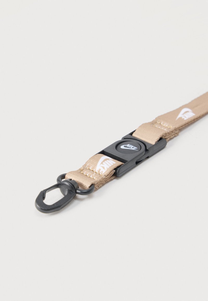 Lanyard beige de Nike con clip de plástico negro y gancho de metal, con logotipos blancos de Nike Swoosh en la tela, colocado plano sobre una superficie clara.