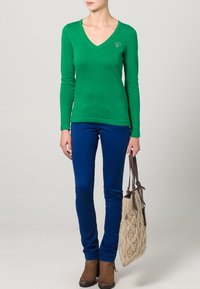 Zelený dlouhý svetr s výstřihem do V v kombinaci s modrými úzkými kalhotami. Hnědé kotníkové boty a béžová pletená kabelka se vzorem.