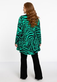 Langärmlige Tunika in Grün mit schwarzen Tigerstreifen, mit abgerundetem Saum und weichem, dehnbarem Stoff; kombiniert mit schwarzen, ausgestellten Hosen.