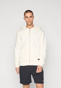 Svijetlo krem zip-up dukserica napravljena od mekanog materijala, s kapuljačom na vezanje, prednjim džepovima i malim logom branda na donjoj strani.