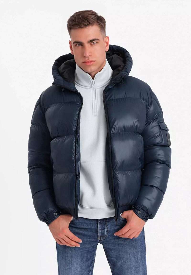 Giacca puffer navy con cappuccio, caratterizzata da una texture trapuntata e una cerniera frontale. Indossata sopra una felpa grigia chiara, abbinata a jeans blu.