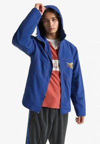 Giacca blu con zip e cappuccio, dotata di tasche anteriori, un grafico colorato e cuciture a contrasto. Indossata sopra una maglietta arancione e pantaloni neri.