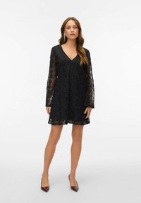 Černé krajkové šaty s dlouhými rukávy a výstřihem do V. Šaty mají květinový vzor a áčkový střih, dosahující délky do poloviny stehen.