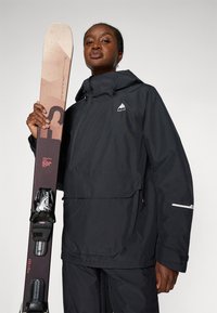 Černá voděodolná bunda s přední kapsou v kombinaci se snowboardem s texturovaným béžovým vrškem a tmavými vázáními, který je držení vztyčený.