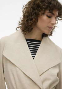 Beige kabát na míru se širokým, strukturovaným límcem, texturovanou látkou a černobílým pruhovaným vrchním topem viditelným pod ním.