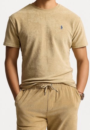 Mann trägt beige Frottee-Kurzarm-T-Shirt und passende Shorts mit Kordelzug, die Hände in den Taschen, am linken Handgelenk eine Armbanduhr.