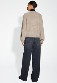 Vrouw met haar in een bun draagt een beige glinsterende trui, een zwarte coltrui en wijde donkere jeans terwijl ze tegenover een witte muur staat.