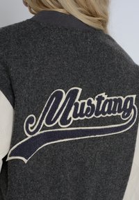 Grå varsityjakke med cremefarvede læderærmer og et stort, broderet "Mustang" logo i marineblå på ryggen.