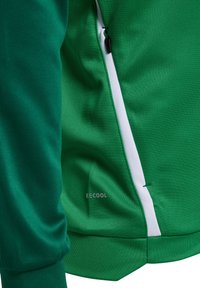 Close-up van een groene jas met een zak met ritssluiting en witte bies en het logo "B:ECOOL" op de stof nabij de zijnnaad.