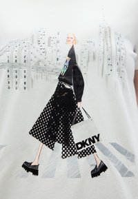 Λευκό μπλουζάκι με γραφικό σχέδιο μιας φιγούρας με μαύρη φούστα με πουά, που κρατά μια λευκή τσάντα DKNY, πάνω σε φόντο πόλης.