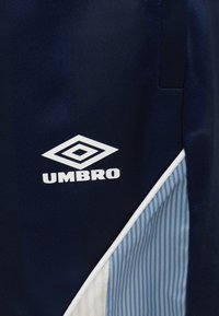 Υφασμα σε ναυτικού μπλε χρώμα με λευκό λογότυπο Umbro και μια διαγώνια περιοχή με κάθετες ρίγες σε ανοιχτό μπλε και λευκό, περιτριγυρισμένη από λευκή μπορντούρα.