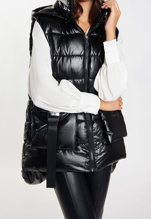 Kvinde iført en skinnende sort polstret vest over en hvid bluse med pufærmer og sorte tætsiddende bukser, stående med armene krydset.