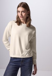 Felpa beige con collo rotondo, polsini a coste e orlo elasticizzato, abbinata a jeans blu scuro con una leggera lucentezza.