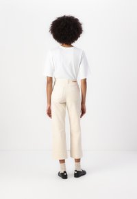 Maglietta bianca a maniche corte abbinata a pantaloni a gamba larga di un leggero color beige. I pantaloni presentano due tasche posteriori e dettagli di cucitura sottili. Indossate scarpe nere.