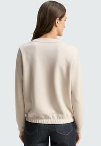 Beżowa bluza z długimi rękawami, zaokrąglonym dekoltem i elastycznym pasem, zestawiona z ciemnymi dżinsami. Gładka faktura i minimalistyczny design.