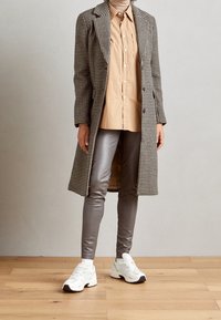 Houndstooth-gepatroneerde jas in zwart en wit, tan kleurige overhemd, metallic grijze leggings en witte sneakers met accenten.