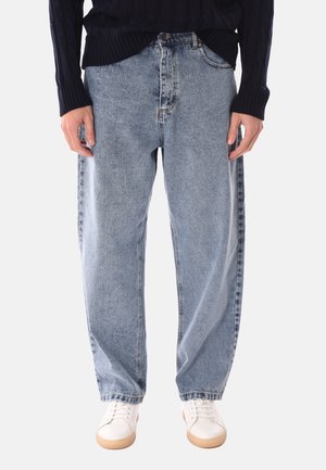 Persona che indossa jeans larghi a gamba larga azzurri, scarpe da ginnastica bianche e un maglione a trecce blu scuro, in piedi con le mani rilassate lungo i fianchi.