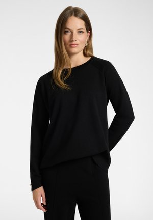 Vrouw met lang lichtbruin haar, gekleed in een zwarte trui met lange mouwen en een zwarte broek, staand tegen een effen witte achtergrond.