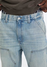 Jeans di denim azzurro chiaro con vita alta, tasche frontali con dettagli di cuciture e chiusura con bottoni. Tessuto strutturato con finitura scolorita.