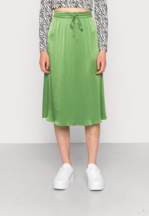 Kobieta nosząca błyszczącą zieloną spódnicę midi z troczkiem w talii, białe sneakersy na platformie oraz długi crop top w nadruk zebry.