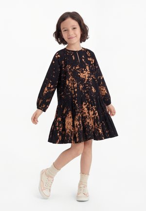 Niño con cabello corto y oscuro que lleva un vestido negro de manga larga con estampado de salpicaduras naranjas, calcetines crema y zapatillas blancas, sonriendo ligeramente.