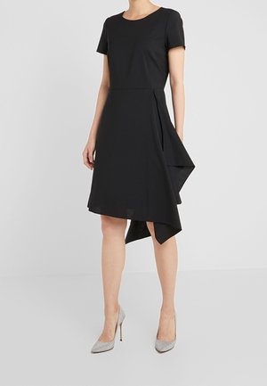 Femme portant une robe noire à manches courtes avec un ourlet asymétrique et des chaussures à talons hauts pailletées argentées sur un fond uni.