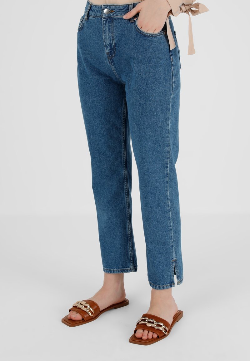 Femme portant un jean bleu taille haute avec de petites fentes latérales et des sandales plates en cuir marron avec des détails en chaîne dorée.