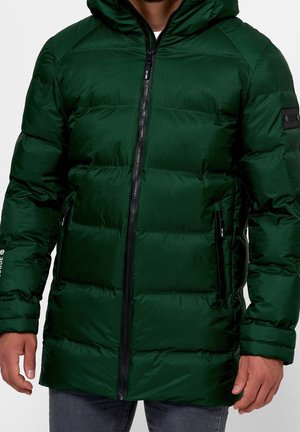 Uomo che indossa una giacca invernale imbottita verde scuro con cappuccio, cerniera frontale e tasche laterali con cerniera, mani rilassate ai lati.