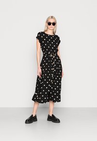 Schwarzes Kleid mit weißen Punkten, kurze Ärmel, Gürtelschnalle in der Taille, geknöpfte Vorderseite, fließender Rock, getragen mit schwarzen Plateauschuhen und Sonnenbrille.