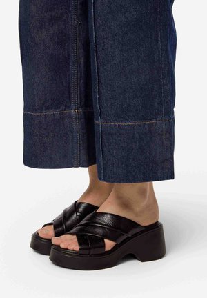Sandali in pelle nera con ampie cinghie intrecciate, suola piatta spessa e dita dei piedi visibili. Abbinati a pantaloni in denim scuri a vita alta e taglio cropped.