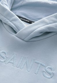 Ljusblå huvtröja med broderad "SAINTS" logotyp, mjukt bomullsmaterial och en avslappnad halsringning. Har en klassisk huva.