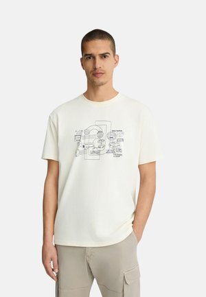 Uomo con capelli corti che indossa una maglietta bianca con un disegno nero a forma di timbro di viaggio e pantaloni cargo beige, in piedi davanti a uno sfondo semplice.