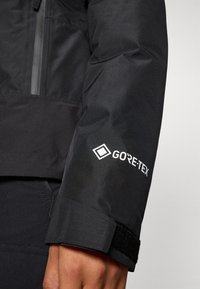 Zwarte waterdichte jas met een gestructureerde stof, klittenbandsluiting bij de manchetten en een wit GORE-TEX-logo op de mouw.