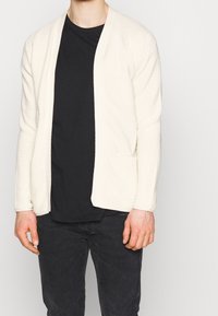 Mann in einer cremefarbenen, gerippten, offen getragenen Strickjacke über einem schwarzen T-Shirt und schwarzen Jeans, der vor einem einfarbigen hellen Hintergrund steht.