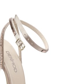 Sandalye con tacco alto beige con finitura in raso, sottile cinturino alla caviglia e dettagli metallici lungo il cinturino. Nome del marchio "DERIMOD" visibile sulla suola.