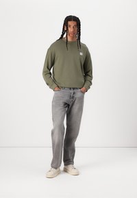 Sweatshirt vert olive à manches longues avec un petit logo, associé à un jean gris clair et des baskets beiges. Textures solides, coupe décontractée.
