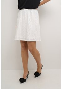 Jupe blanche, de longueur genou avec des détails en œillets, taille élastique et coupe ample. Portée avec des chaussures à talons ouverts noires ornées de roses.