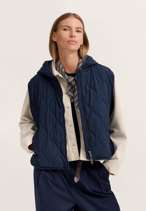 Frau trägt eine marineblaue gesteppte Kapuzenweste über einer beigen Jacke und einem schwarz-weiß gestreiften Schal, steht mit den Händen in den Taschen.