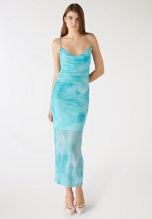 Giovane donna in piedi che indossa un vestito lungo aderente senza maniche, tinto a nodi blu, e scarpe beige con punta appuntita e fiocchi.