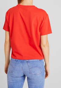 T-shirt in cotone rosso con maniche corte, colletto rotondo e vestibilità rilassata, abbinata a jeans in denim azzurro chiaro con tasche posteriori e un leggero lavaggio.