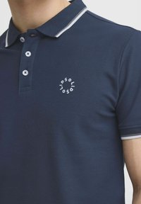 Námornícky polo tričko s textúrovaným povrchom, s trojicou gombíkov a s kolieskovým bielym logom. Obsahuje biele a šedé akcenty na golieri.