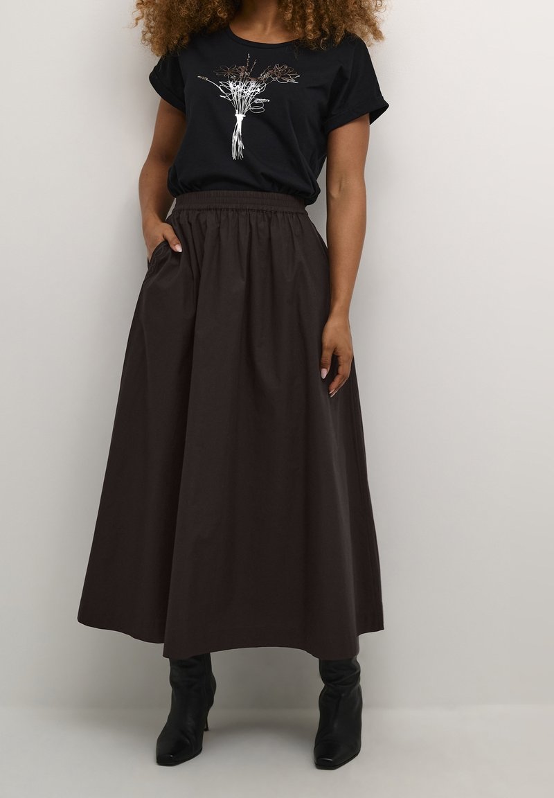 Femme portant un t-shirt noir avec un motif floral blanc rentré dans une jupe longue marron foncé taille haute et des bottines noires.
