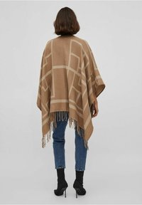 Brązowy ponczo z geometrycznymi wzorami w jaśniejszych odcieniach, z frędzlami na brzegach, noszony na niebieskich dżinsach i czarnych butach na wysokim obcasie.
