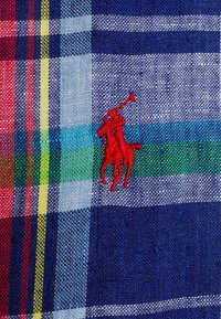 Tessuto a quadri blu e rosso con accenti verdi e gialli. Presenta tessiture intricate e un logo di cavallo ricamato in rosso.