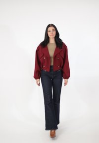 Bordowa krótka kurtka z podwójnym zapięciem na guziki, szerokimi rękawami i dwoma przednimi kieszeniami, zestawiona z dopasowanym beżowym topem i rozszerzanymi ciemnymi dżinsami.