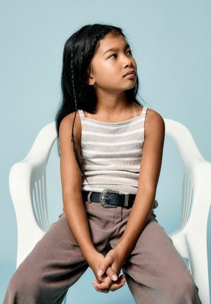 Mladé dievča s dlhými čiernymi vlasmi, oblečená v pruhovanom tielku a hnedých nohaviciach, sediac na bielom plastovom kresle a pozerajúc sa doprava.