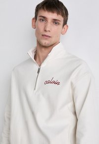 Sudadera de forro polar blanca con cremallera de cuarto y el logo bordado en rojo de "calvin" en el pecho, que presenta una tela texturizada y un cuello alto.