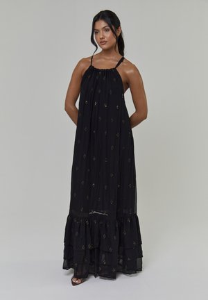 Vrouw die met handen achter de rug staat, gekleed in een mouwloze zwarte maxi-jurk met gouden stippen en een geruffelde zoom, tegen een effen achtergrond.