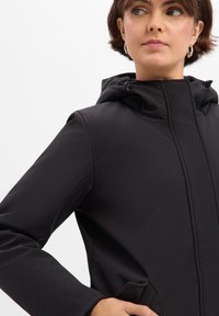 Veste à capuche noire avec une texture lisse, dotée d'une fermeture éclair et de poches latérales. Le design comprend des détails de couture subtils.